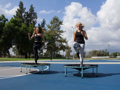 two women jumping on rebounders on a basketball court outside with trees in the background