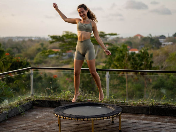 Person bouncing on trampoline outdoors, surrounded by greenery and glass railing.