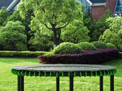 A trampoline sits on green grass with trees and building.