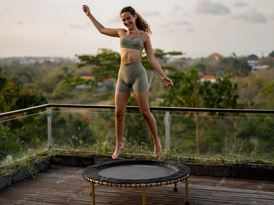 Person bouncing on trampoline outdoors, surrounded by greenery and glass railing.